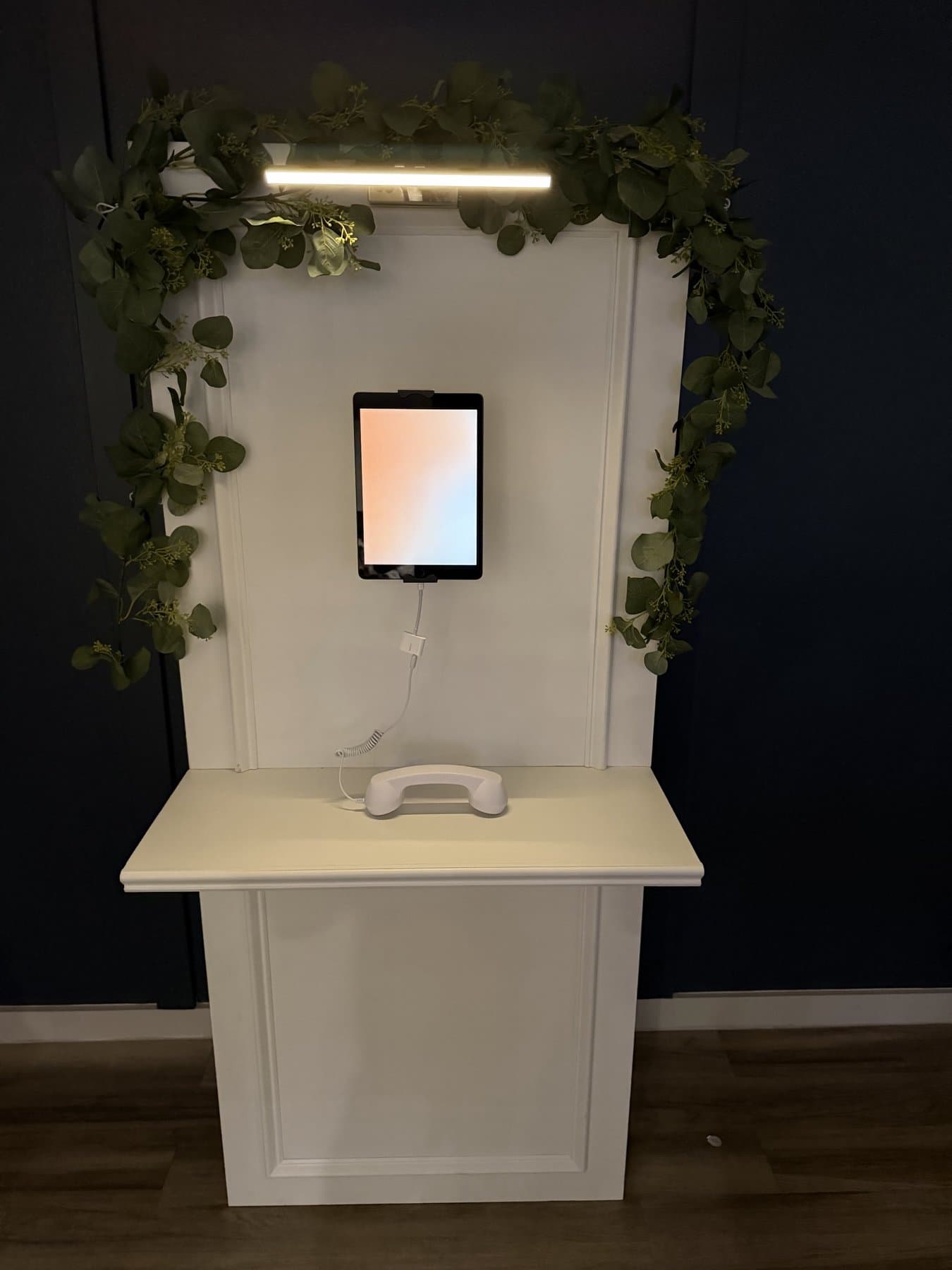 The open-air Memory Wall — a cream paneled wall with a brass picture light, iPad recording screen and white handset, wrapped in eucalyptus garland against a navy wall